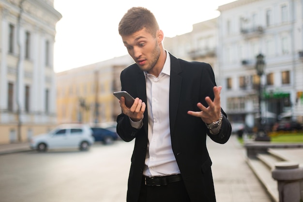 Jonge zakenman in zwarte jas en wit overhemd die verbaasd op mobiel kijkt terwijl hij tijd doorbrengt op straat met prachtig uitzicht op de stad op de achtergrond