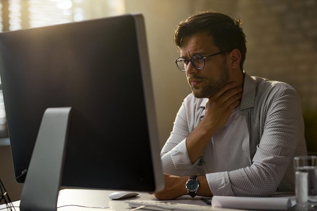 Jonge zakenman voelt pijn in zijn keel tijdens het werken op een desktop-pc op kantoor