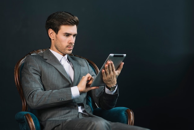 Jonge zakenmanzitting in leunstoel die digitale tablet gebruiken tegen donkere achtergrond