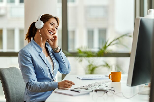 Jonge zakenvrouw die plezier heeft en muziek luistert via een koptelefoon terwijl ze op een computer op kantoor werkt