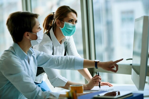 Jonge zakenvrouw en haar collega die gezichtsmaskers dragen terwijl ze op een computer op kantoor werken tijdens een virusepidemie