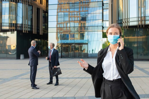 Jonge zakenvrouw in gezichtsmasker en kantoor pak praten op mobiele telefoon buitenshuis. Ondernemers en stadsgebouwen op achtergrond. Kopieer ruimte. Bedrijfs- en epidemisch concept