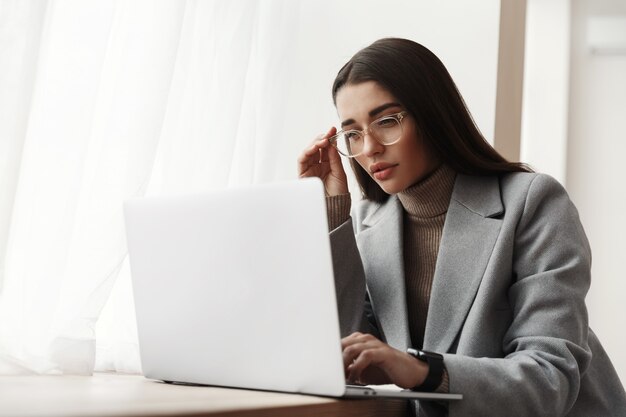 Jonge zakenvrouw in glazen zitten in een kantoorgebouw, werken op een laptop.