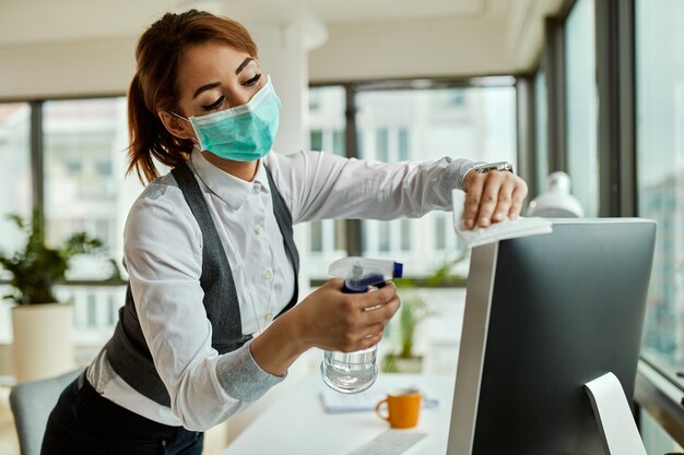 Jonge zakenvrouw met gezichtsmasker die haar desktop-pc desinfecteert terwijl ze op kantoor werkt tijdens de coronavirusepidemie