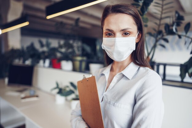 Jonge zakenvrouw met gezichtsmasker die op kantoor staat en klembord met documenten vasthoudt