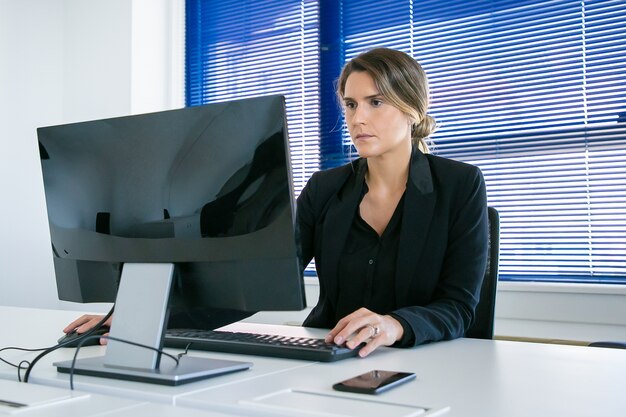 Jonge zakenvrouw werken in haar kantoor, met behulp van computer op de werkplek, kijken naar display gericht. Gemiddeld schot. Digitale communicatie of bedrijfsleidersconcept