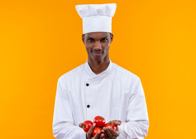 Jonge zelfverzekerde Afro-Amerikaanse kok in uniform chef houdt aardappelen met beide handen geïsoleerd op een oranje achtergrond met kopie ruimte