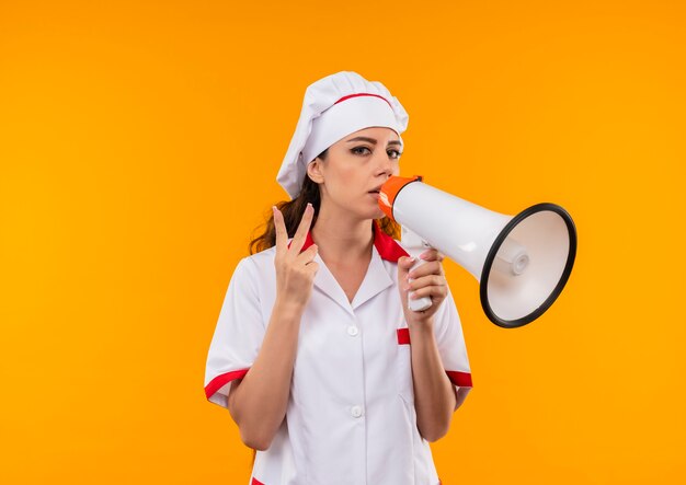 Jonge zelfverzekerde blanke kok meisje in uniform chef houdt luidspreker en gebaren overwinning handteken geïsoleerd op oranje muur met kopie ruimte