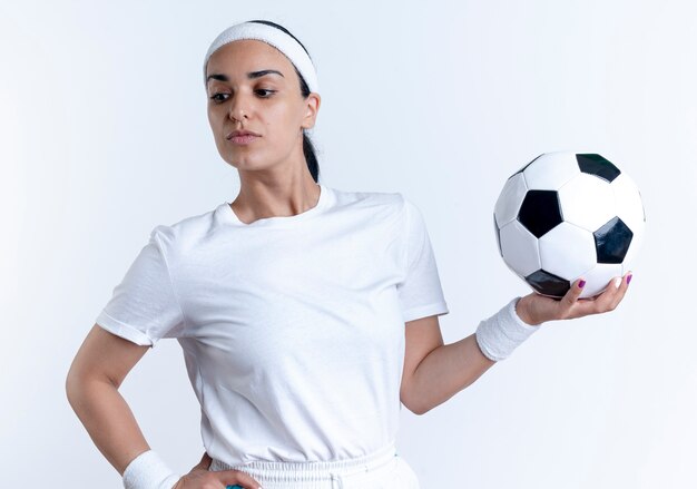 jonge zelfverzekerde blanke sportieve vrouw met hoofdband en polsbandjes houdt bal naar kant kijken