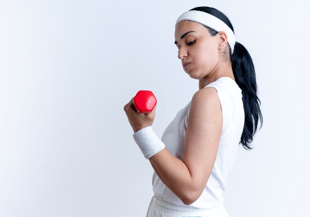 Jonge zelfverzekerde blanke sportieve vrouw met hoofdband en polsbandjes oefeningen en kijkt naar halter geïsoleerd op witte ruimte met kopie ruimte