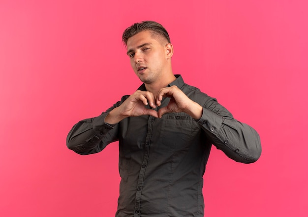 Gratis foto jonge zelfverzekerde blonde knappe man gebaren hart hand teken kijken naar camera geïsoleerd op roze achtergrond met kopie ruimte