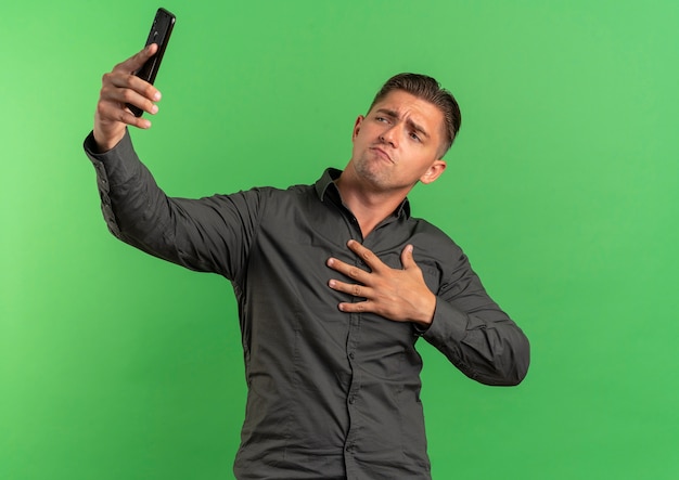 Jonge zelfverzekerde blonde knappe man legt hand op de borst kijkt naar telefoon nemen selfie geïsoleerd op groene ruimte met kopie ruimte