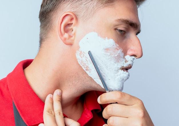 Jonge zelfverzekerde blonde mannelijke kapper in uniform besmeurd gezicht met scheerschuim scheren met scheermes geïsoleerd op witte ruimte met kopie ruimte