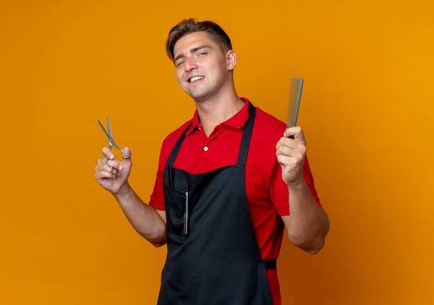 Jonge zelfverzekerde blonde mannelijke kapper in uniform houdt kam en schaar geïsoleerd op oranje ruimte met kopie ruimte