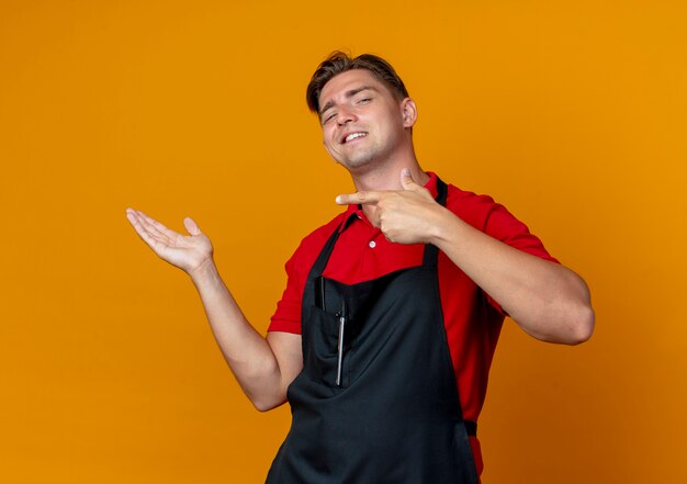 Jonge zelfverzekerde blonde mannelijke kapper in uniforme punten op lege hand geïsoleerd op oranje ruimte met kopie ruimte