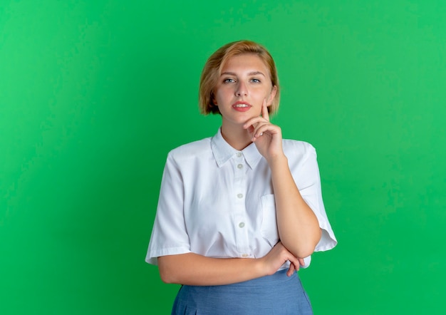Jonge zelfverzekerde blonde Russische meisje legt hand op kin kijken camera geïsoleerd op groene achtergrond met kopie ruimte