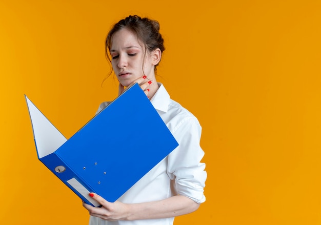 Jonge zelfverzekerde blonde Russische meisje legt hand op kin kijken naar bestandsmap op oranje met kopie ruimte