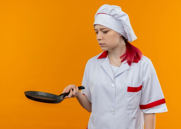 Jonge zelfverzekerde blonde vrouwelijke chef-kok in eenvormige chef-kok houdt en kijkt naar koekenpan geïsoleerd op oranje muur