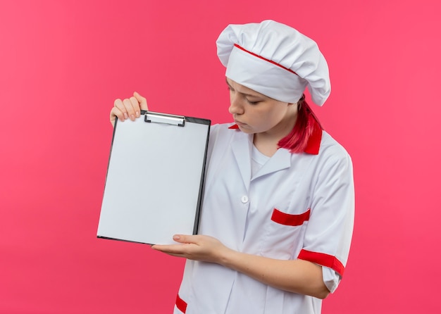 Jonge zelfverzekerde blonde vrouwelijke chef-kok in uniform chef-kok houdt en kijkt naar klembord geïsoleerd op roze muur
