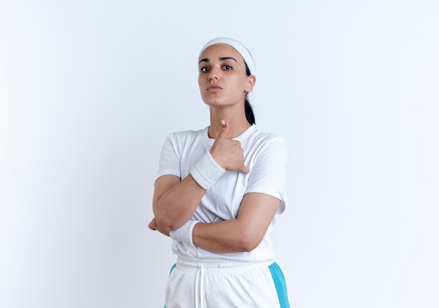 Jonge zelfverzekerde Kaukasische sportieve vrouw die hoofdband en polsbandjes draagt, kruist armen en duimen omhoog geïsoleerd op witte ruimte met kopie ruimte