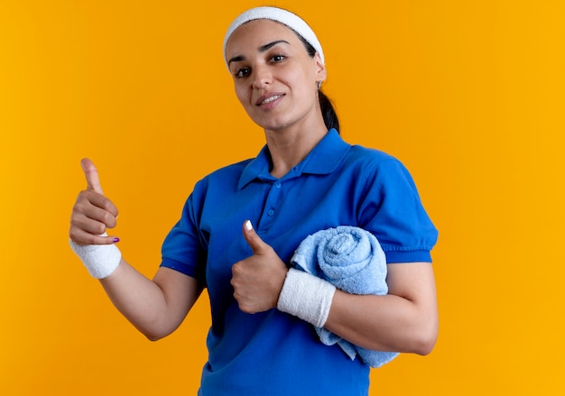 Jonge zelfverzekerde Kaukasische sportieve vrouw met hoofdband en polsbandjes beduimelt omhoog handdoek met arm op oranje met kopie ruimte te houden