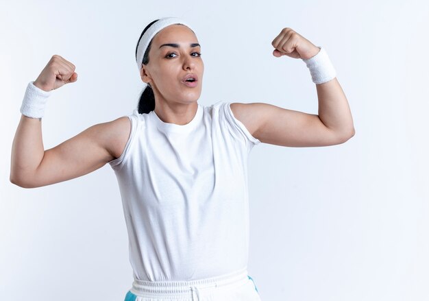 Jonge zelfverzekerde Kaukasische sportieve vrouw met hoofdband en polsbandjes gespannen biceps geïsoleerd op een witte ruimte met kopie ruimte