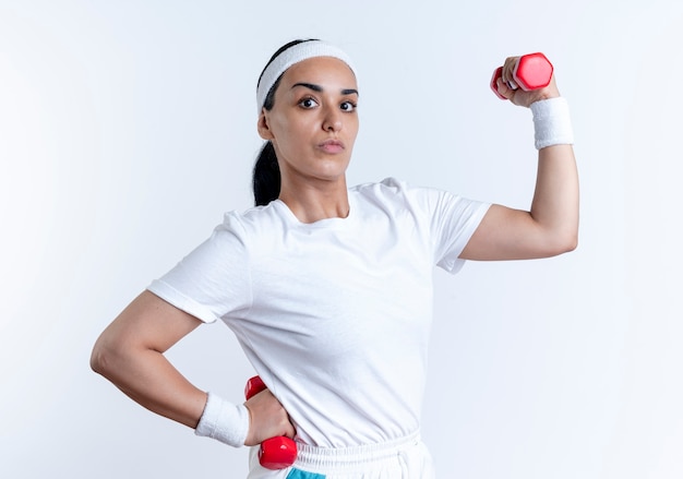 Jonge zelfverzekerde Kaukasische sportieve vrouw met hoofdband en polsbandjes gespannen biceps houden halters geïsoleerd op witte ruimte met kopie ruimte