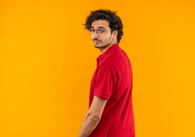 Jonge zelfverzekerde man in rood shirt met optische bril staat met rug en kijkt geïsoleerd op oranje muur