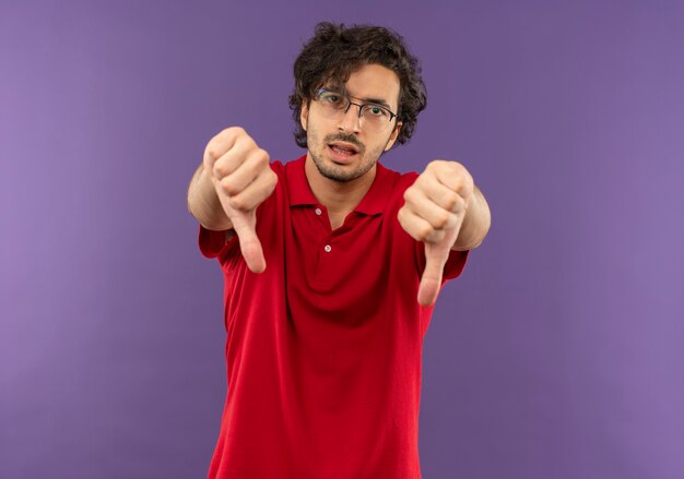 Jonge zelfverzekerde man in rood shirt met optische bril thumbs down geïsoleerd op violette muur