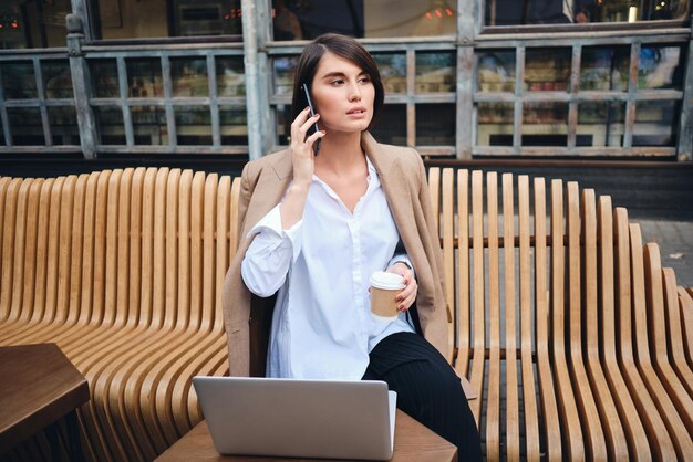 Jonge zelfverzekerde stijlvolle zakenvrouw met laptop praten op mobiele telefoon tijdens koffiepauze in café op straat