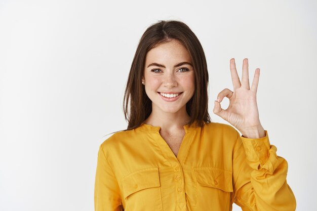 Jonge zelfverzekerde vrouw die lacht, een goed gebaar toont, ja of goed zegt en op een witte muur staat