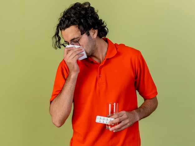 Jonge zieke man met bril met glas water en pak medische tabletten zijn neus afvegen met servet geïsoleerd op olijfgroene muur met kopie ruimte