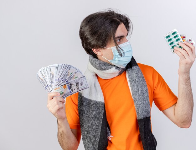 Jonge zieke man met sjaal en medisch masker contant geld vasthouden en kijken naar pillen in zijn hand geïsoleerd op wit