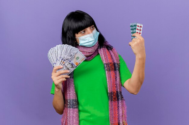 Jonge zieke vrouw die masker en sjaal draagt die geld en verpakkingen van capsules houdt die capsules bekijken die op purpere muur met exemplaarruimte worden geïsoleerd