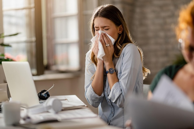 Jonge zieke zakenvrouw niest in een tissue terwijl ze op kantoor werkt