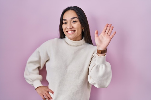 Gratis foto jonge zuid-aziatische vrouw die over een roze achtergrond staat en afziet van hallo, gelukkig en glimlachend vriendelijk welkomstgebaar