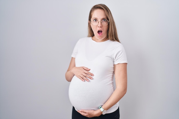 Jonge zwangere vrouw die een baby verwacht, zwangere buik bang en geschokt aanraakt van verbazing en verbaasde uitdrukking, angst en opgewonden gezicht.