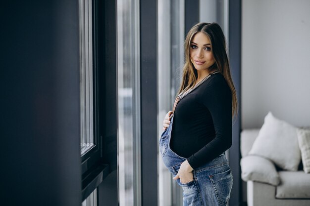 Jonge zwangere vrouw die haar buik vasthoudt en bij het raam staat