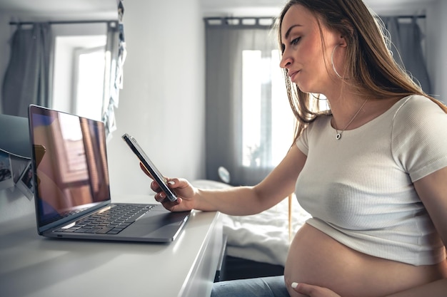 Jonge zwangere vrouw gebruikt thuis smartphone en laptop