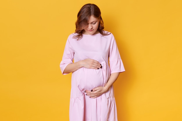 jonge zwangere vrouw met bruin haar, in elegante roze jurk staande met handen op haar buik voor geel, neerkijkend op haar buik, heeft donkere manicure.