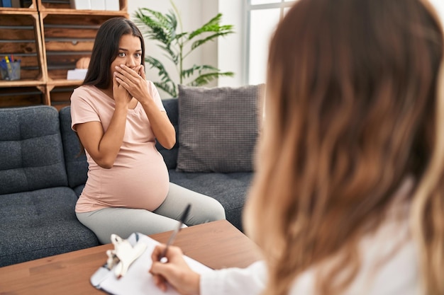 Jonge zwangere vrouw tijdens therapiesessie geschokt mond bedekkend met handen voor fout. geheim begrip.
