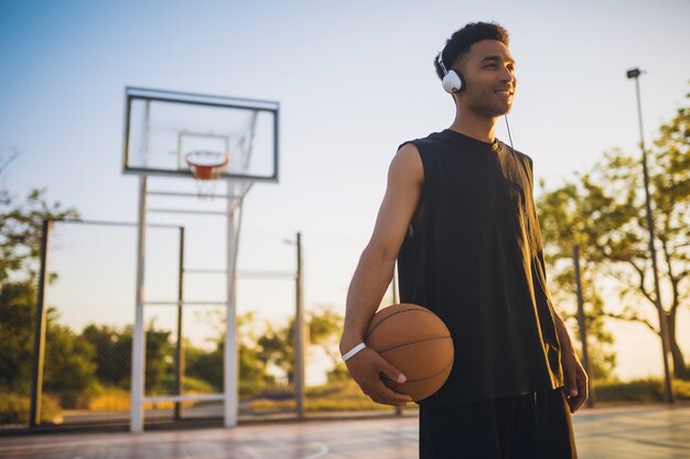 Jonge zwarte man sport doen, basketbal spelen op zonsopgang, luisteren naar muziek op koptelefoon, actieve levensstijl, zomerochtend