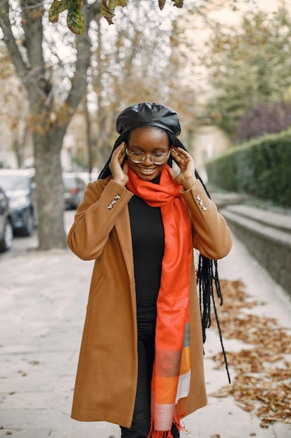 Jonge zwarte vrouw met lange locs-haarstijlen die buiten staan. Vrouw met bruine jas, oranje sjaal en zwarte hoed