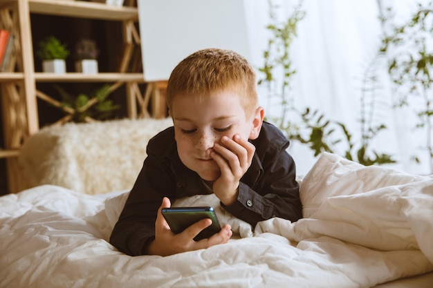 Jongen die thuis verschillende gadgets gebruikt. klein model met slimme horloges, smartphone of tablet en koptelefoon. selfie maken, chatten, gamen, video's bekijken. interactie van kinderen en moderne technologieën.