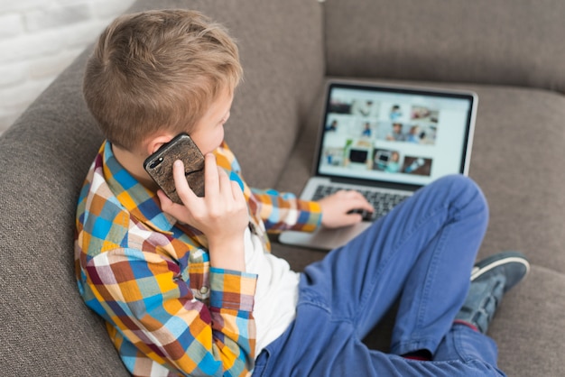 Gratis foto jongen met behulp van laptop op de bank en het maken van telefoongesprek