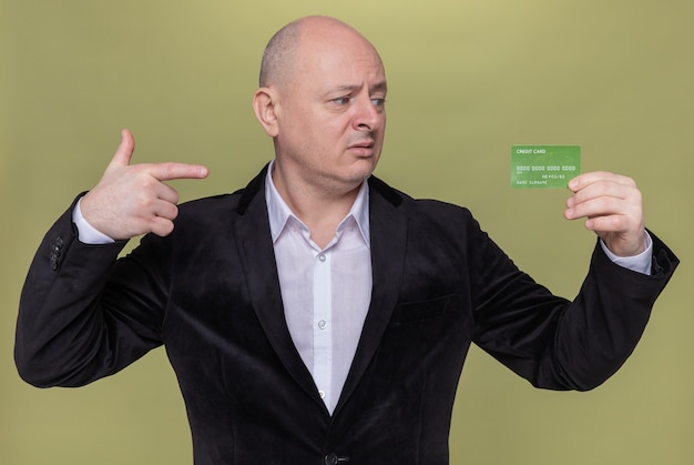 Gratis foto kale man op middelbare leeftijd in de creditcard die van de kostuumholding met wijsvinger richt met het verwarren uitdrukking die zich over groene muur bevindt