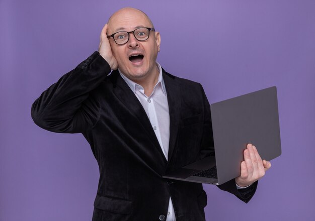 Kale man van middelbare leeftijd in pak met bril die laptop vasthoudt en opzij kijkt, verward met de hand op zijn hoofd voor een fout die over de paarse muur staat