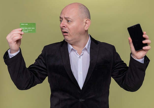 Kale man van middelbare leeftijd in pak met smartphone en creditcard te kijken naar het verward en erg angstig staande over groene muur