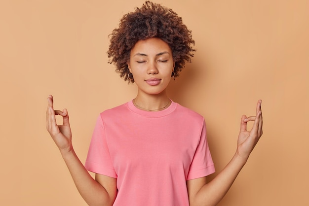 Gratis foto kalme geconcentreerde jonge vrouw houdt ogen gesloten vingers bij elkaar mediteert binnen beoefent yoga draagt casual roze t-shirt geïsoleerd over bruine achtergrond probeert vrede te vinden staat in zen pose