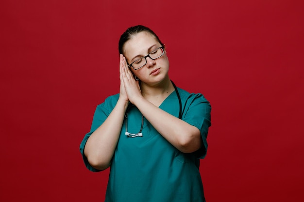 Kalme jonge vrouwelijke arts met een uniforme bril en een stethoscoop om haar nek die slaapgebaar maakt met gesloten ogen geïsoleerd op rode achtergrond